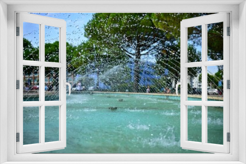 Fototapeta Naklejka Na Ścianę Okno 3D - View of tiny drops of water gushing from a fountain. Blue water surface and many small drops of flying water. View into the stream inside the fountain. Background with defocused green trees, blue sky.
