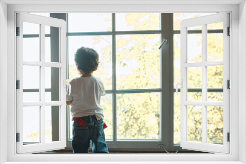 Indoor rear view of little boy with brown curly hair standing on windowsill by large window, looking outside, spending day at home alone, feeling bored. Childhood, family and domesticity concept