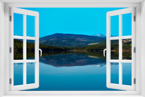Fototapeta Naklejka Na Ścianę Okno 3D - Calm lake surrounded by wild green pine forests and snowy mountain ranges in the distance.