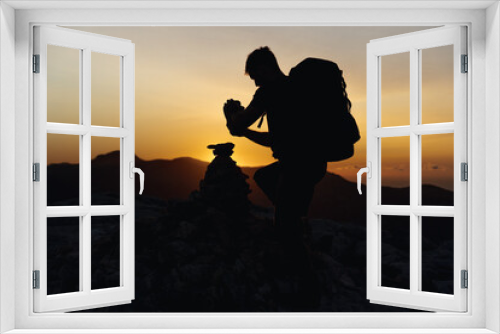 Fototapeta Naklejka Na Ścianę Okno 3D - Hiker at the top of a mountain stacking stones in a small hill with a beautiful golden hour sunset illuminating the silhouette, carrying a large backpack