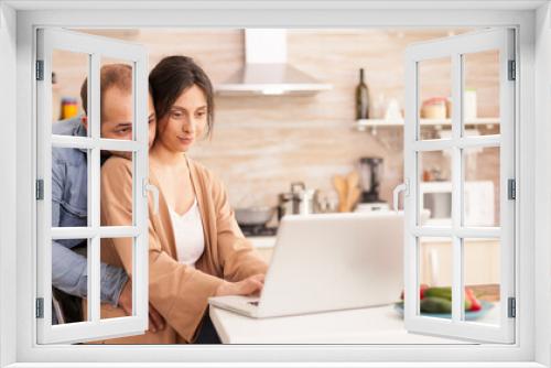 Woman using laptop in kitchen while her husband hugs her. Happy loving cheerful romantic in love couple at home using modern wifi wireless internet technology