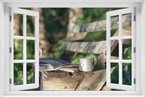 Fototapeta Naklejka Na Ścianę Okno 3D - A book with a mug of tea on a wooden bench in the forest. The background is blurred.