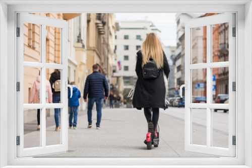 Rear view of girl riding public rental electric scooter in urban city environment at fall. New eco-friendly modern public city transport in Ljubljana.