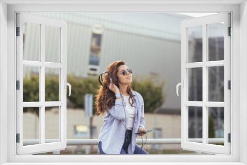 Young pretty student girl, waiting for a taxi car, and listening to music from the phone on big headphones