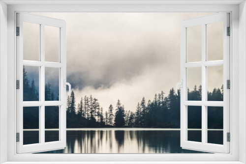 Fototapeta Naklejka Na Ścianę Okno 3D - Silhouettes of pointy tree tops on hillside along mountain lake in dense fog. Reflex of pines to calm water of highland lake. Alpine tranquil landscape at early morning. Ghostly atmospheric scenery.