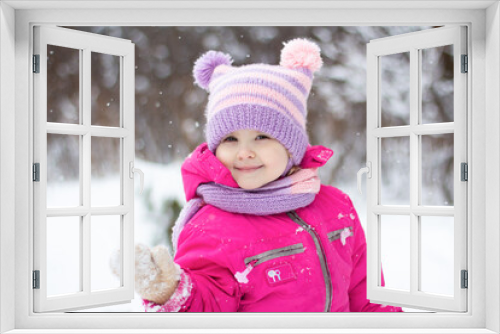 Smiling little girl waving her hand with snow saying hello on a winter day. Beautiful snowy portrait of a cute girl wearing bright pink jacket, knit cap with pompons and scarf. Selective focus.