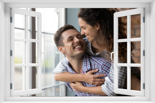 Enjoying one another. Happy attractive millennial couple of spouses lovers having romantic date at home smiling embracing by window. Affectionate young husband holding beloved laughing wife on back