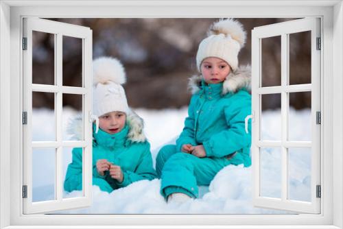 Two funny little girls in identical clothes are having fun in a beautiful winter park. Cute kids playing in the snow. Winter activities for children.