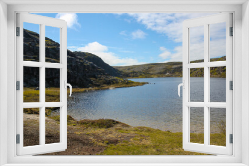 Fototapeta Naklejka Na Ścianę Okno 3D - The lake is located in the mountains. Green-yellow grass surrounds the lake. Beautiful summer landscape on a sunny day. The stone hills are illuminated by the sun. White clouds on a bright blue sky.