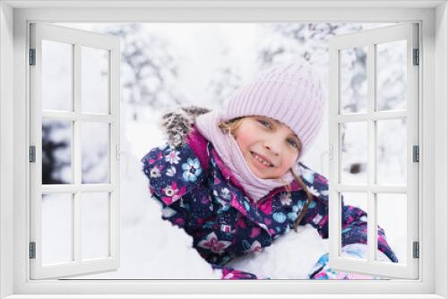 Little smiling girl, kid walking in beautiful winter forest, park among trees, branches covered with snow. Pink hat, scarf, snood, bright colorful warm jacket. Blue eyes, blond hair.Fashionable image