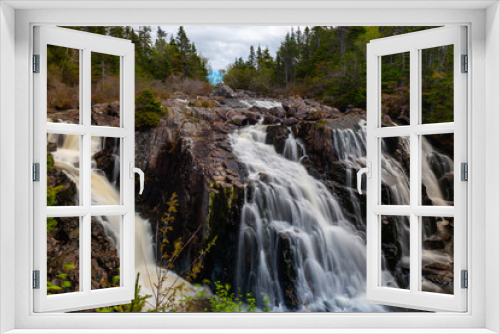 Fototapeta Naklejka Na Ścianę Okno 3D - A raging river of white rapids and waterfalls with tall evergreen trees on both sides. The stream is enclosed by large boulders or rock formations with dead read leaves and moss covering greenery. 