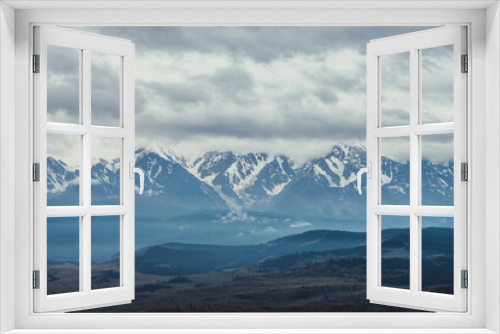 Fototapeta Naklejka Na Ścianę Okno 3D - Scenic mountain landscape with great snowy mountain range among low clouds and green forest in valley at early morning. Atmospheric alpine scenery with blue white high mountain ridge under cloudy sky.