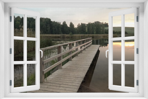 Fototapeta Naklejka Na Ścianę Okno 3D - A wooden footbridge by the lake in wild nature landscape. A recreation area scenic view in the evening at sunset time. The calm surface of the water reflects sunlight. Grassy lakeshore a scenic view.