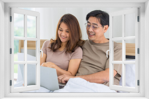 happy middle age Asian couple dressed casually sitting relaxed on a bed at home enjoy looking at a laptop computer. Warmth, delightful family concept