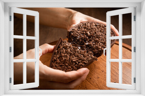 Fototapeta Naklejka Na Ścianę Okno 3D - Woman hands crack a stuffed chocolate easter egg with grated chocolate on the top on a wooden stand on a wooden table.