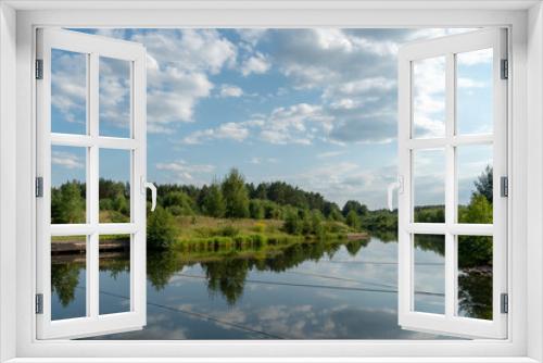 Fototapeta Naklejka Na Ścianę Okno 3D - Calm on the river and the reflection of the sky and clouds in the water. Canal and ferry crossing. Ropes are stretched across the river. The banks of the river are overgrown with bushes and trees.