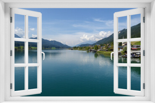 Fototapeta Naklejka Na Ścianę Okno 3D - An idyllic, panoramic view on the Weissensee lake in Austria. The lake is surrounded by high Alps. There is a small village at the lake's shore, with tall church tower. Few clouds. Calmness and peace
