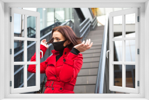 A young woman wearing a black protective medical mask near a glass building with a staircase in a red coat. Coronavirus epidemic, protective measures, new reality