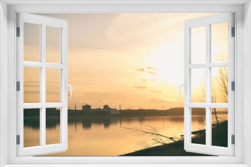 Fototapeta Naklejka Na Ścianę Okno 3D - Scenic view of a beautiful sunset over the river in the city in spring against the background of the golden sky, the sun over the water and silhouettes of bushes in the foreground. Spring landscape.