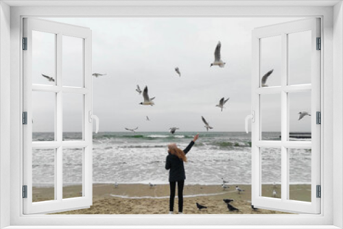 Fototapeta Naklejka Na Ścianę Okno 3D - A girl with long hair standing on the beach and feeding seagulls with bread with a beautiful sea background. Girl feeds seagulls and pigeons at the seashore. 