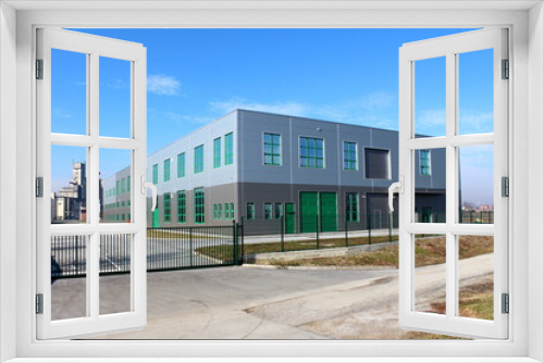 Fototapeta Naklejka Na Ścianę Okno 3D - Newly built modern grey and green production industrial building surrounded with closed metal fence and large old concrete storage silo building in background on warm sunny winter day