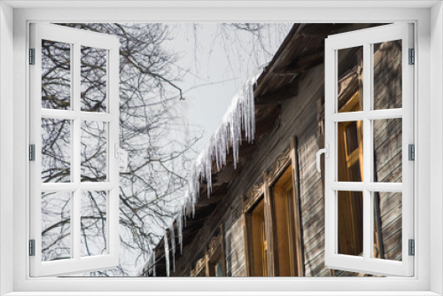 Fototapeta Naklejka Na Ścianę Okno 3D - Icicles hang from the roof of a wooden house and shine in the sun. Against the background of a gray sky, branches of a tall tree. Abnormal weather concept. New Year's and Christmas is over.
