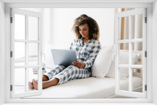 The smiling black woman sitting on the bed in front of laptop near window