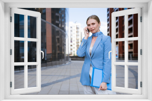 caucasian business woman in a blue jacket and dress talking on the phone with a folder of papers in her hand against the background of an office building