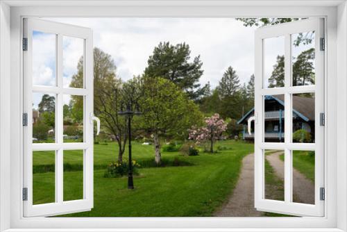 Fototapeta Naklejka Na Ścianę Okno 3D - Old two story wooden house by the road. Countryside. Villa cottage made of dark wood with large terrace and balcony. Garden design with flowering trees and daffodils. Green lawn is mowed. Lantern.