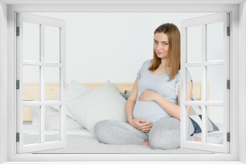 Pregnant young European woman sitting on a large bed and stroking her big belly, 7 month pregnancy, waiting for the birth of her baby.