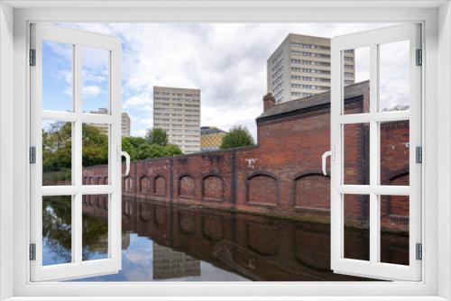 Fototapeta Naklejka Na Ścianę Okno 3D - Row of 1960s and 1970s residential tower block flats and skyscrapers behind red brick wall reflecting in Birmingham canal waterways. Cramped living conditions in high rise block,