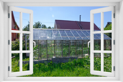 Fototapeta Naklejka Na Ścianę Okno 3D - Roofs of wooden houses and greenhouses in the village against the background of blue sky and green foliage. Hot sunny summer day. Medium plan