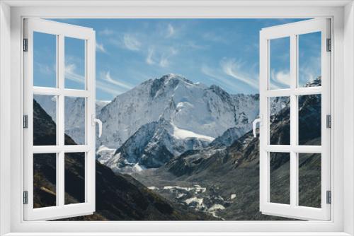 Fototapeta Naklejka Na Ścianę Okno 3D - Alpine sunny landscape with high snowy mountain with peaked top and glacier between black rocks under cirrus clouds in sky. Big snow covered mountains in sunshine. White-snow pointy peak in sunlight.
