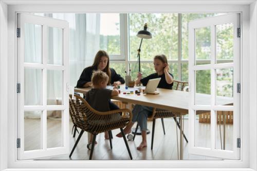  mom and her children girls are sitting at the table and drawing with paints using an online lesson on a tablet           