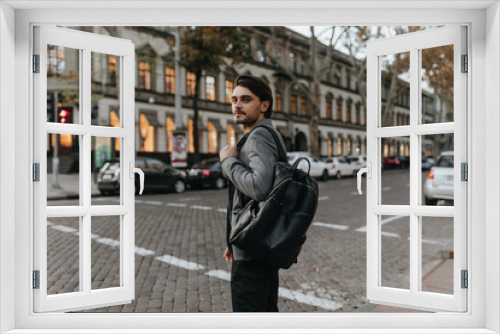 Lifestyle portrait of charming young brunette in grey blazer and black pants, looking into camera and holding backpack. Man posing against day city background