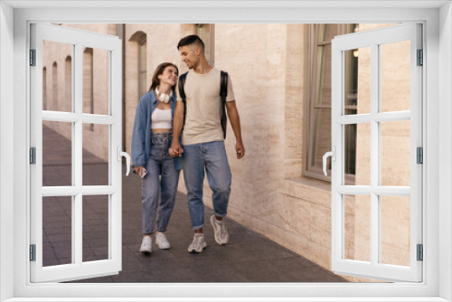 Full-length photo of young couple walking on street. Pretty dark-haired lady wearing denim outfit and white sneakers smiling and holding hand boyfriend in jeans and t-shirt