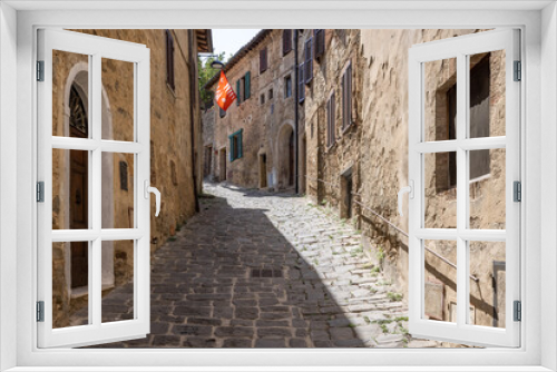 Fototapeta Naklejka Na Ścianę Okno 3D - A narrow medieval stone paved street with a red waving flag in the tuscanian town of Montalcino