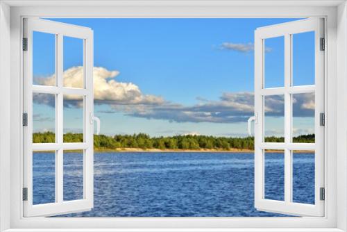 Fototapeta Naklejka Na Ścianę Okno 3D - Young green forest along the sandy shore on blurred background of blue lake and blue sky with white clouds