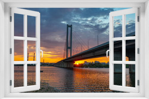 Fototapeta Naklejka Na Ścianę Okno 3D - South bridge in Kiev. Sunset over the Dnieper. Thick clouds over the evening city. Evening shot of the bridge. Orange sun at sunset. The rays break through the clouds and are reflected in the river.