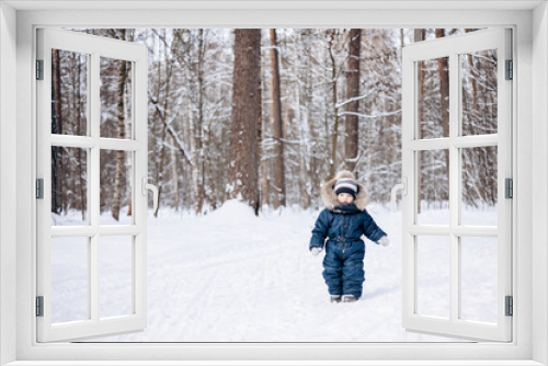 Child walking in snowy spruce forest. Little kid boy having fun outdoors in winter nature. Christmas holiday. Cute toddler boy in blue overalls and knitted scarf and cap playing in park.