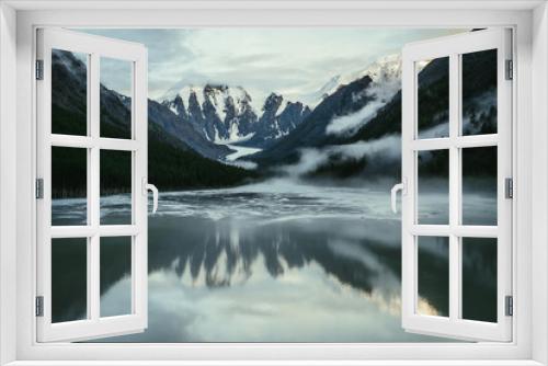 Fototapeta Naklejka Na Ścianę Okno 3D - Scenic alpine landscape with snowy mountains in golden sunlight reflected on mirror mountain lake in fog among low clouds. Atmospheric highland scenery with low clouds on rocks and green mirror lake.