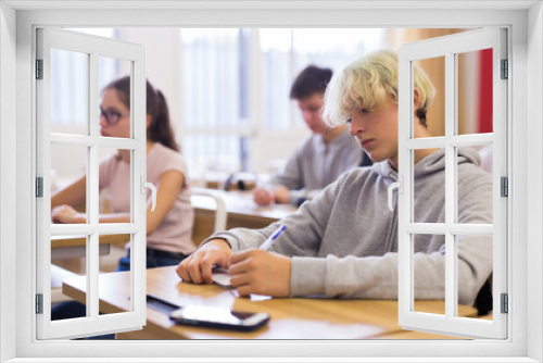 Smart teenager studying in classroom, listening to lecturer and writing in notebook..