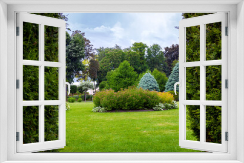 Fototapeta Naklejka Na Ścianę Okno 3D - landscape desing of a park with a garden bed and trees with leaves and pine needles on a green lawn, evergreen and seasonal plants in the backyard look through thuja border.