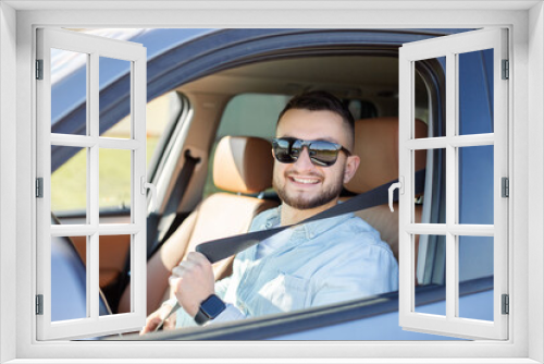 Portrait of young man sitting in driving seat of car, fastening safety belt.