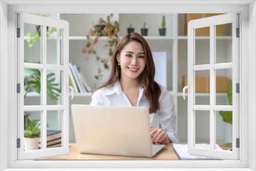 Successful smiling beautiful young Asian businesswoman sitting with laptop and computer while doing some paperwork at the office. Looking at camera.