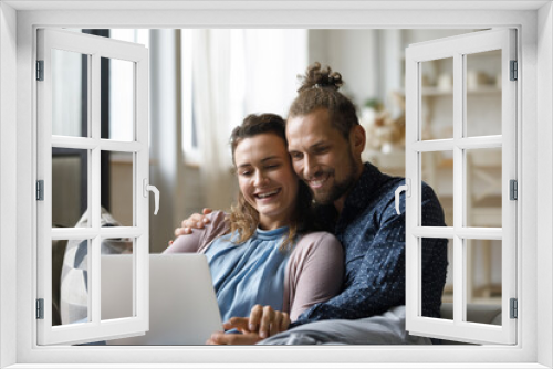 Cheerful happy family couple watching movie on laptop in living room, resting on comfortable couch with computer, using online app, internet service, laughing, talking on video call together