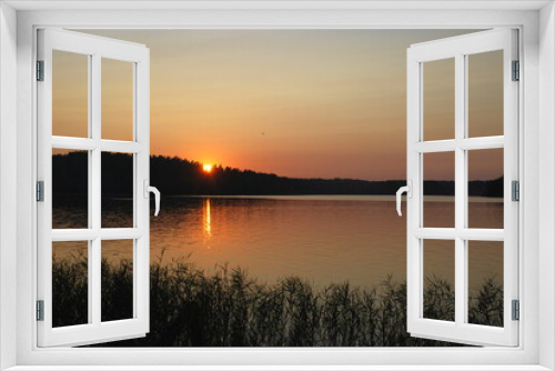 Fototapeta Naklejka Na Ścianę Okno 3D - Orange sunset on the shore of a forest lake. The sun went down low over the forest growing on the shore of the lake. The surface of the water is almost smooth, the sky is clear without clouds.