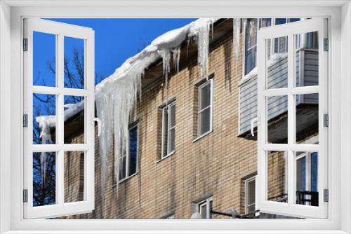Fototapeta Naklejka Na Ścianę Okno 3D - Big beautiful icicles hang from the snow-covered roof in front of the windows of the house. Snow on the roof. Frosty sunny day