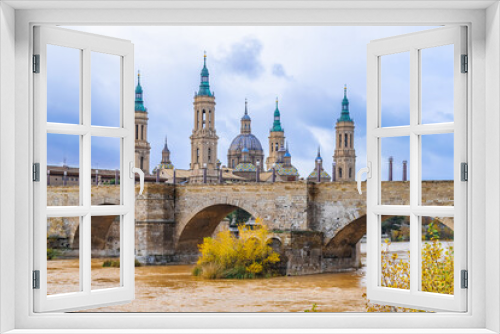 Medieval stone bridge with arches on the backdrop of the towers of Church of Our Lady of the Pillar in Zaragoza, Spain. Cityscape with ancient architecture and yellow water of Ebro river