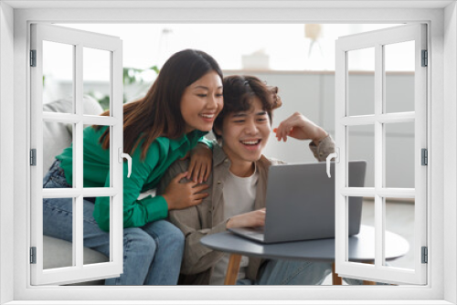 Millennial Asian couple in casual wear using laptop, having online meeting, watching interesting movie in living room